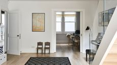 A bright basement space with wooden flooring, a black rug, two small wooden chairs, a piano and storage seen to the left. The staircase running down into the space has a glass balustrade. Through an open door, a desk space with wooden chairs with fluffy throws is seen.