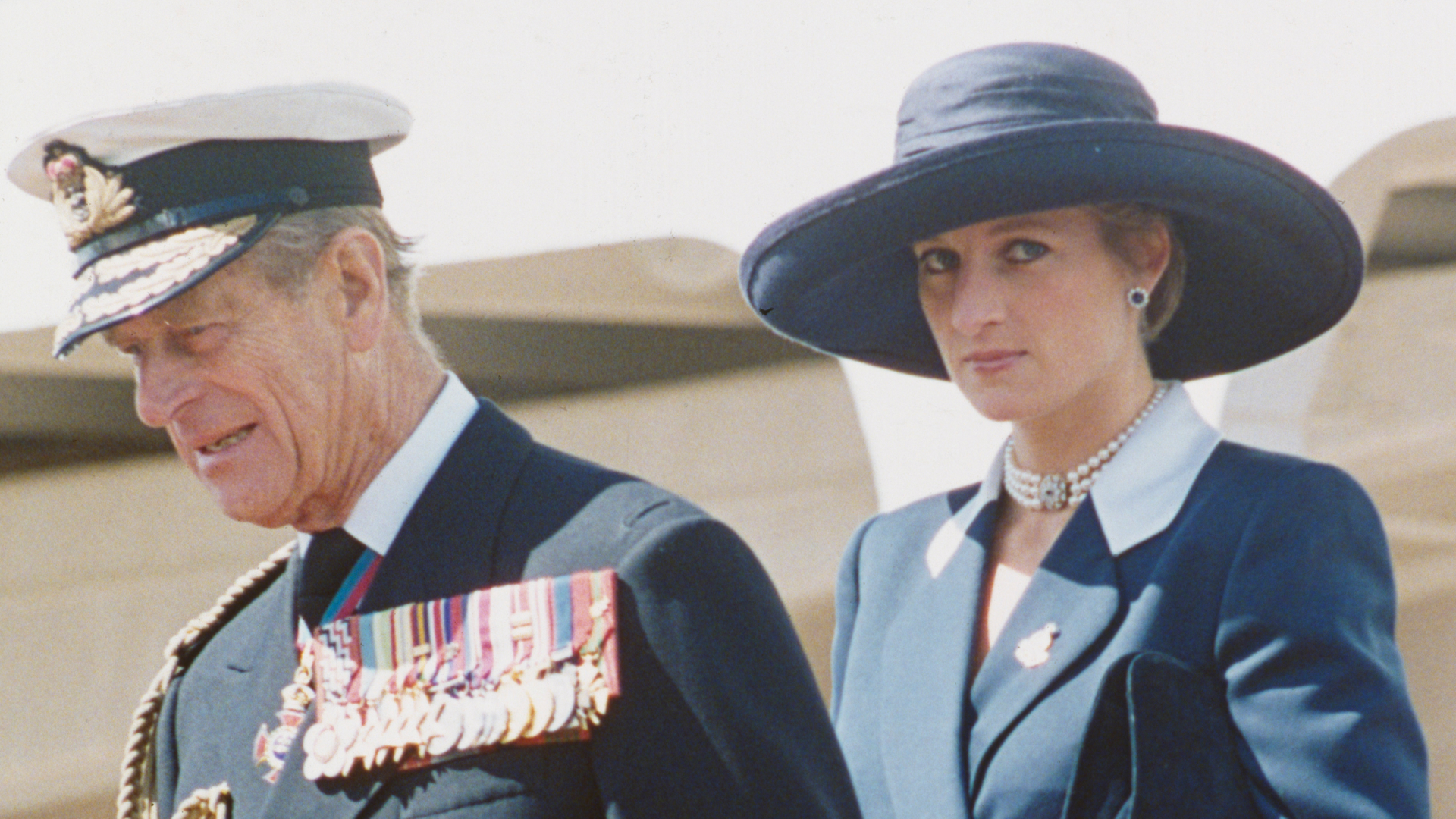 Prince Philip wears military uniform and Princess Diana wears a navy coat dress with a large-brim hat