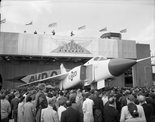 a jet plane with pointy nose in front of a large crowd of people. the photo is in black and white given the age