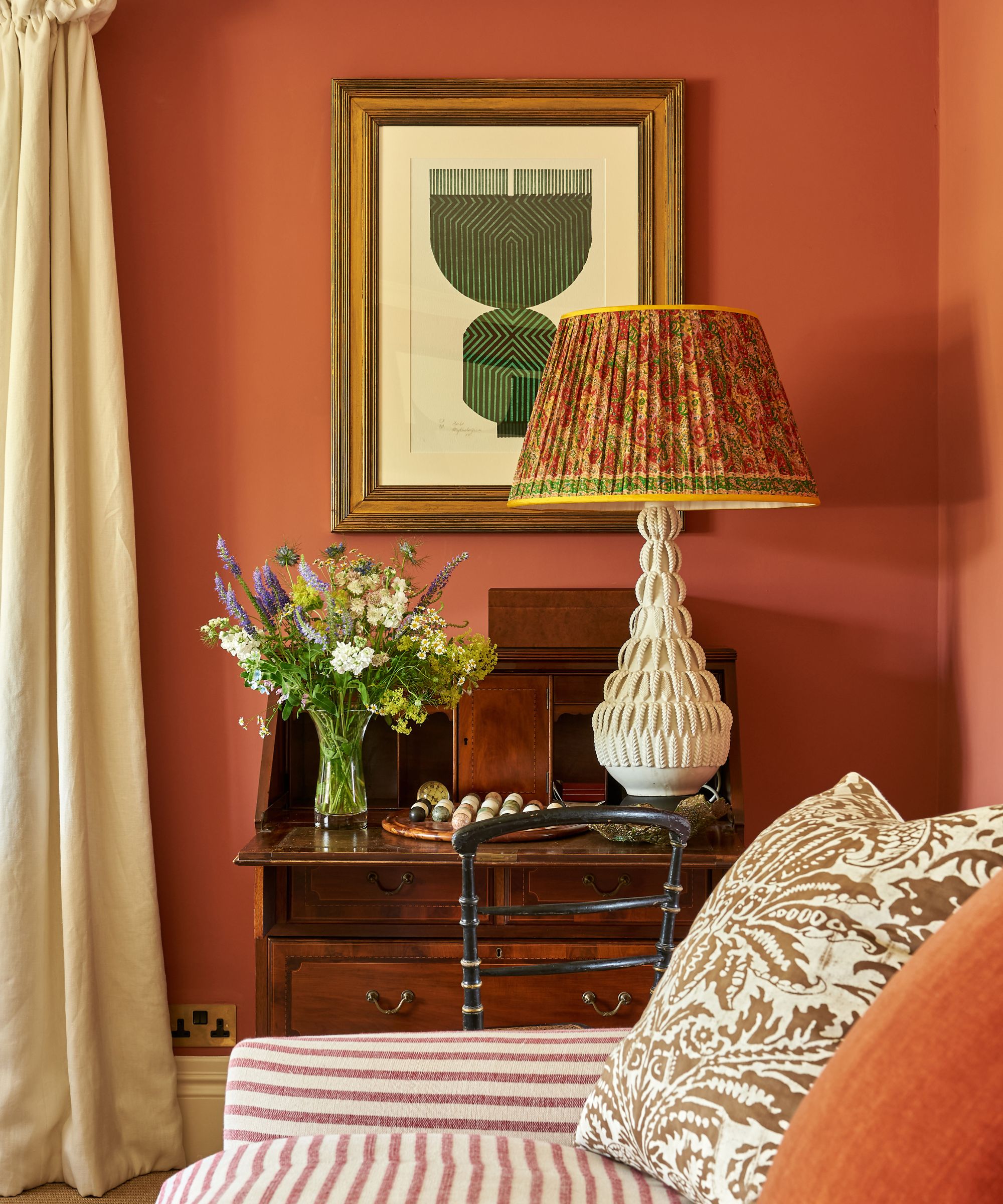 Living room with earthy red painted walls, cream drapes, mahogany writing desk, striped sofa, floral cushion and large table lamp with floral pleated shade and vase of flowers