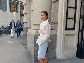 Woman wears white sheer long-sleeve button down and long denim shorts.