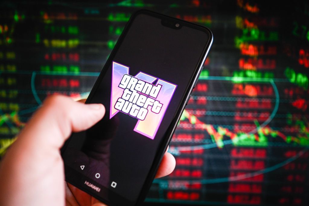 Grand Theft Auto (GTA) VI logo is displayed on a smartphone with stock market percentages in the background.