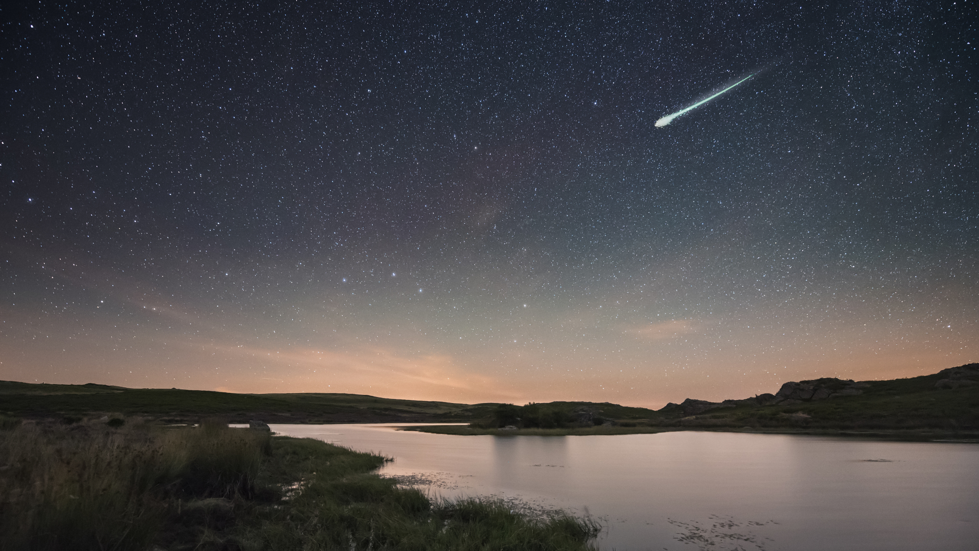 A bright meteor is pictured streaking through a starry sky above a lake. The glow of dawn is visible close to the horizon.