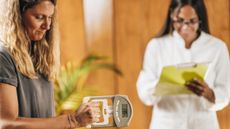 Woman performs grip strength test with digital hand dynamometer with another woman in a white coat and clipboard standing in the background