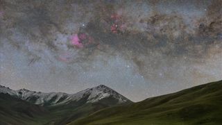 snow-capped mountains are dwarfed by a star-studded night sky with a huge emission nebula glowing red pink.