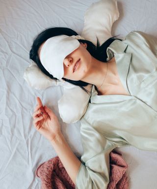 A woman wearing a white sleep mask with a white pillow behind her, wearing light green silk pajamas, lying on white sheets