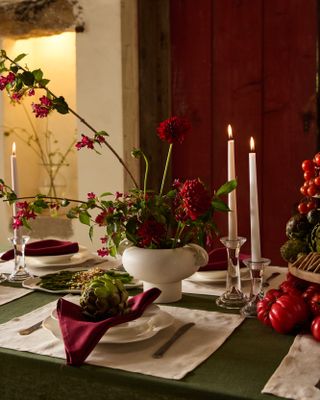 Christmas table setting with a green table cloth, white placemats, white pates, and red napkins. There is an artichoke in the center of the plate, red flower arrangements and white candles burning on the table.