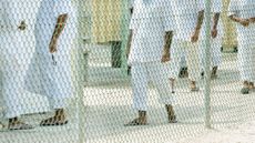 profile shot of detainees at Guantanamo Bay through a chainlink fence. They are dressed in white linen pants and sandals and white lined shirts.