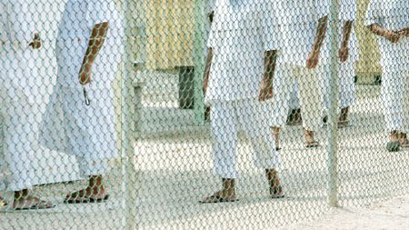 profile shot of detainees at Guantanamo Bay through a chainlink fence. They are dressed in white linen pants and sandals and white lined shirts.