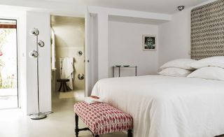Interior view of a room at House of Birds in Portugal featuring white walls, a bed with pillows and white linen, a red and white wooden bench at the end of the bed, framed wall art, a silver floor lamp and partial view of the bathroom