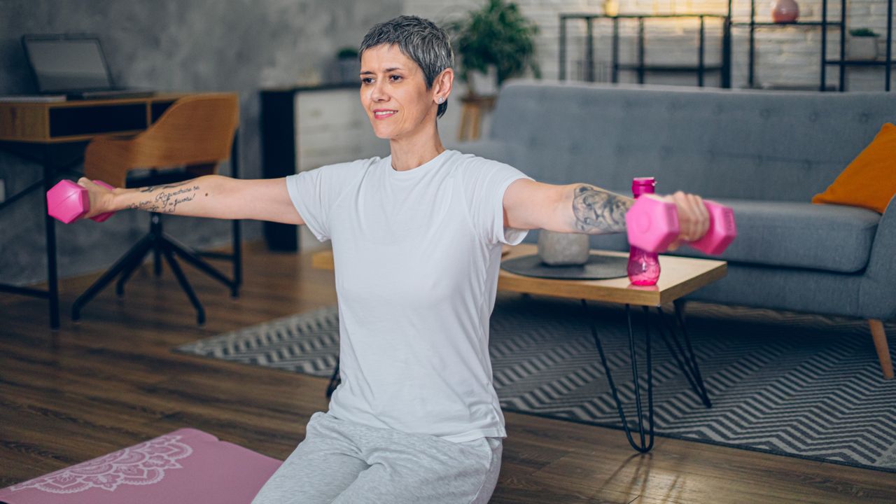 A woman performs a dumbbell kneeling lateral raise in a living room on a yoga mat. Her legs are bent underneath her and her arms are raised out to the sides, a dumbbell grasped in each hand. She is smiling and behind her we see a grey couch, wooden coffee table, a desk and chair and black shelving. 