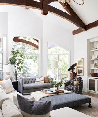 Large living room with white painted walls, dark wood beams on ceiling, large windows, large cream sofa, square striped dark gray ottoman, gray two-seater sofa, black and cream patterned armchair, two framed pictures on wall, alcove shelving in corner