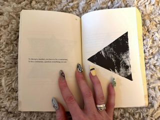 A hand whose nails have been decorated with eccentric nail art in grey, yellow, black, and three-dimensional motifs lays atop an open book whose pages show a quote and a triangular print in black.