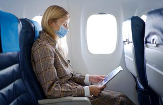 Portrait of young woman with long blond hair wearing mask inside airplane while reading safety instructions.