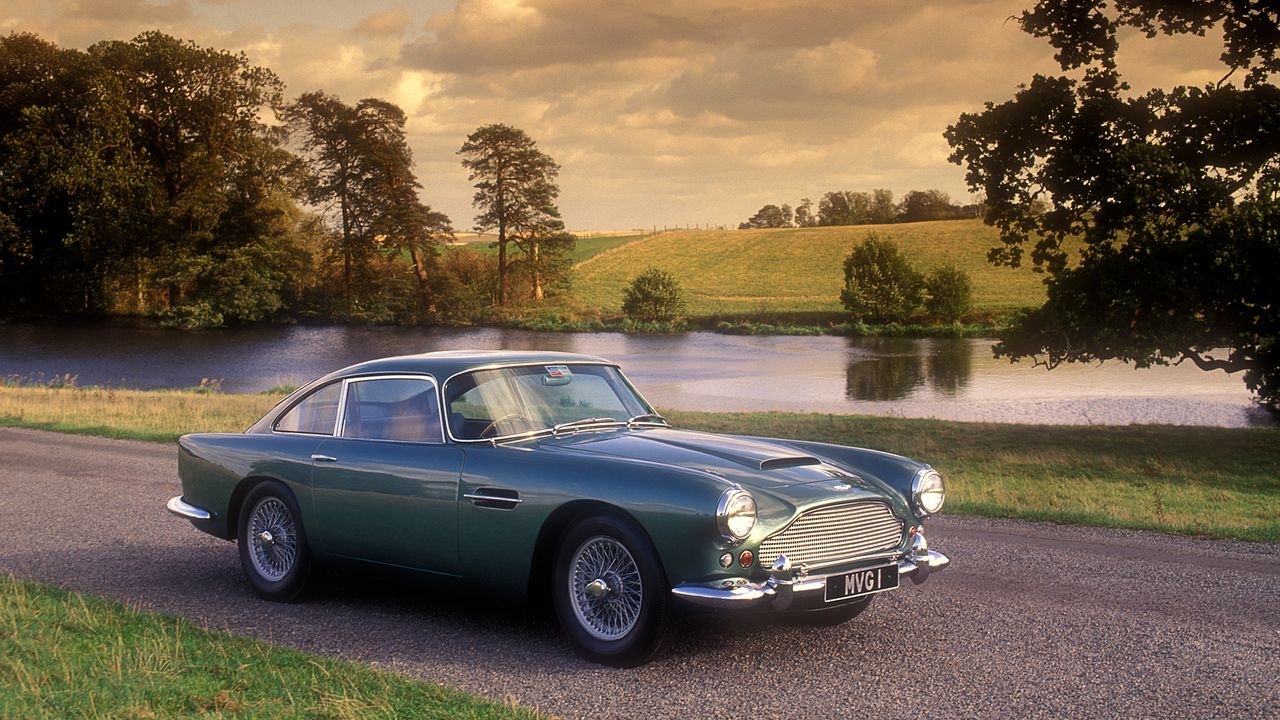 A vintage car on a road passing a body of water with a hill in the background