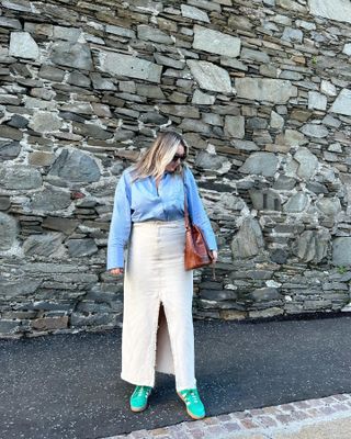 woman in front of stone wall wearing blue button down shirt, white denim maxi skirt, green Adidas sneakers, carrying brown shoulder bag
