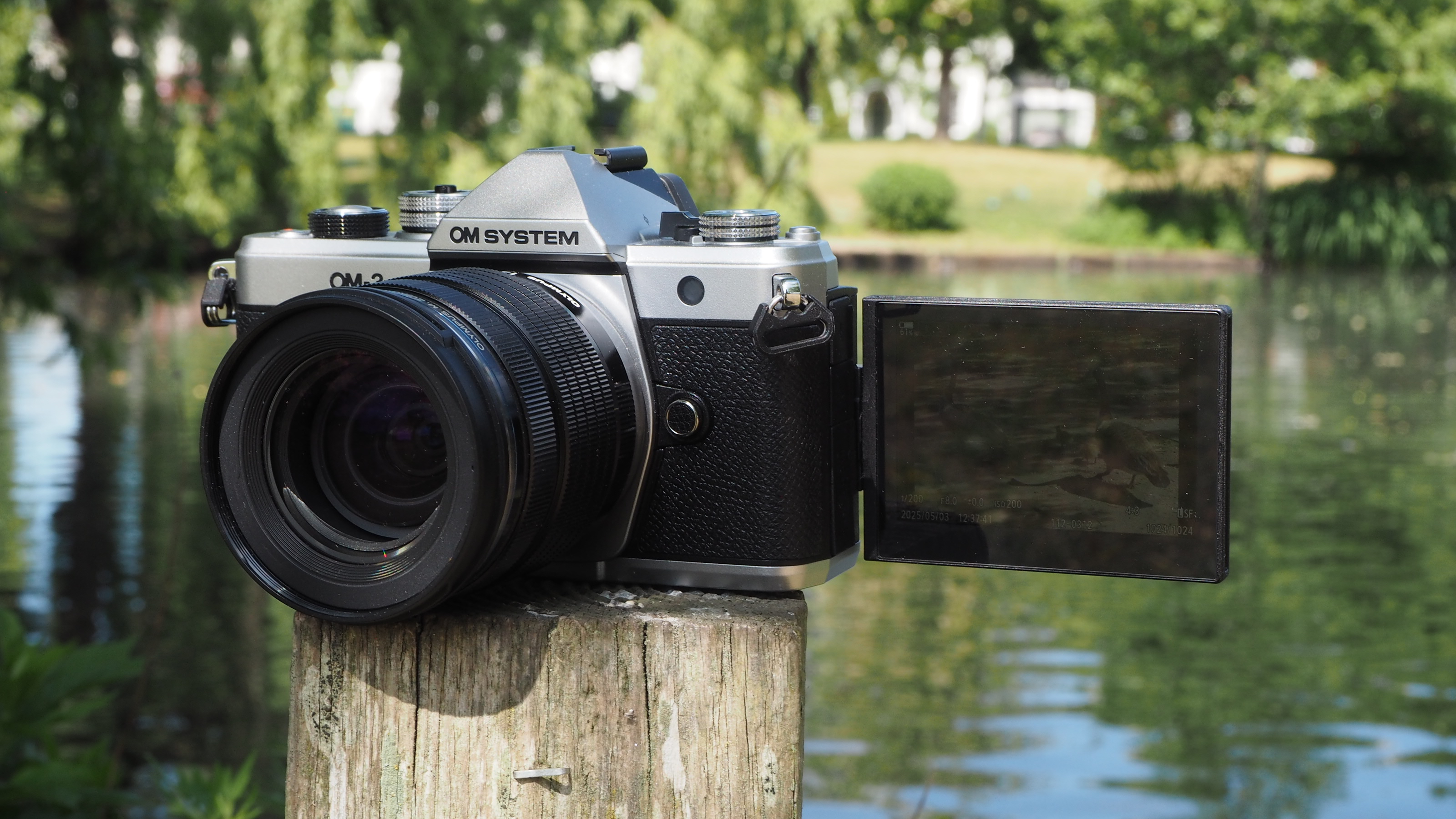 OM System OM-3 with the screen flipped out on a wooden post next to a pond