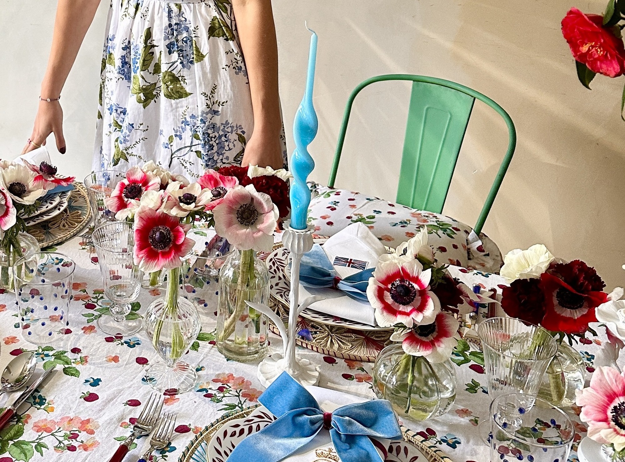 A bright table setting with a floral patterned tablecloth and matching flower centrepieces in small glass vases. The place settings are decorated with a blue velvet bow, and the plates are floral printed.