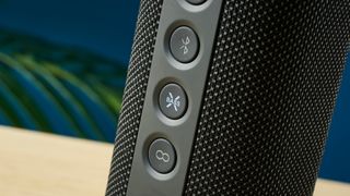 a black oblong-shaped bluetooth speaker with mesh covering and a carry strap photographed on a beige table in front of a navy wall