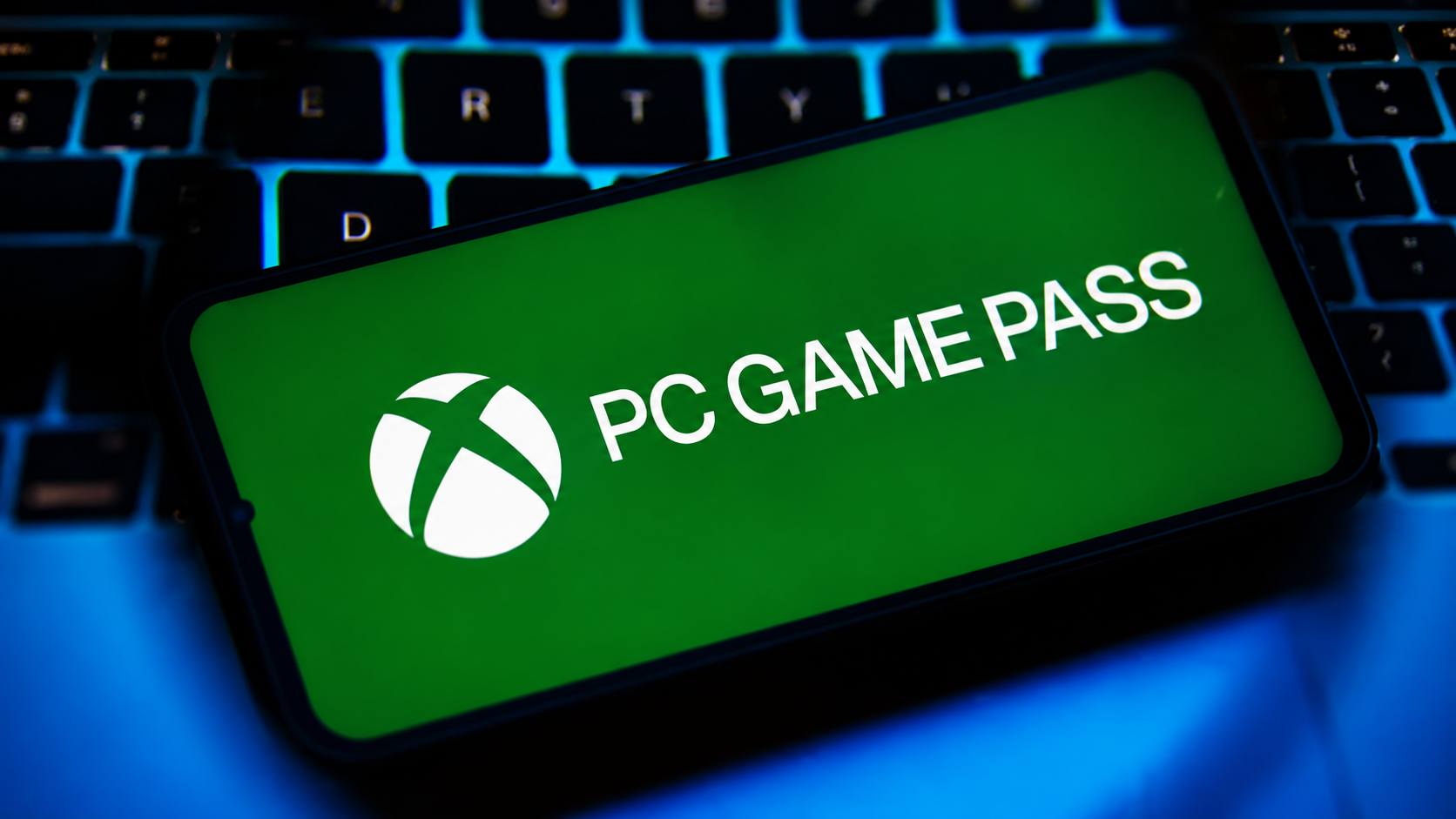 Android smartphone displaying the Xbox PC Game Pass logo on a green screen, resting on a wooden desk in front of an RGB gaming keyboard.