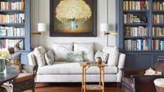 Colorful living room with white sofa and cushions, small wooden cofee table, two armchairs, and large blue bookshelves full of books. A large flower painting is pictured on the wall.