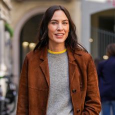Alexa Chung wearing a Miu Miu brown suede jacket with a grey sweater over a yellow T-shirt, baggy jeans, and ballet flats in Milan
