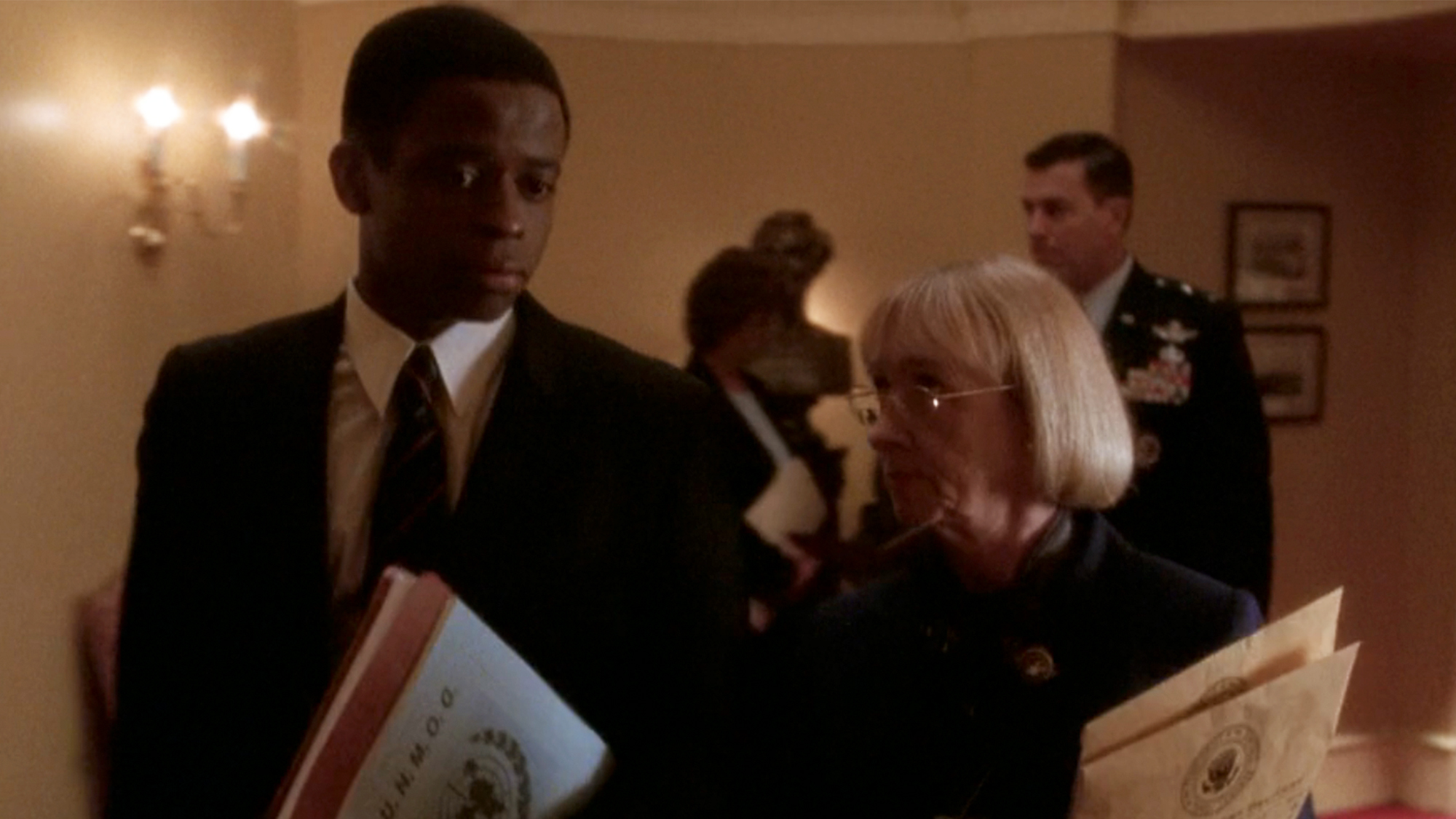Dule Hill in a suit, walking and talking with Mrs. Landingham as she looks at him with disapproval.