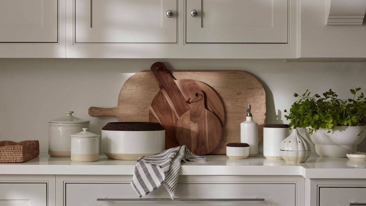 A white kitchen with cutting boards leaning against the back splash, and an array of neutral colored stone wear jars lined up around them. 