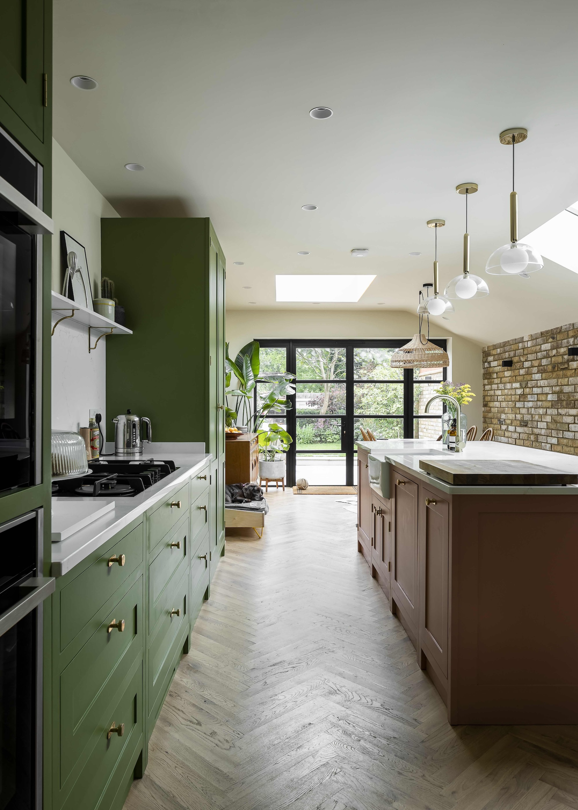 A view of the green kitchen cupboards and terracotta colored island and marble worktops, with herringbone wood flooring and exposed brick wall to the right.
