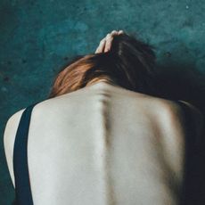 Woman doubled over forwards holding her hair up as seen from behind.