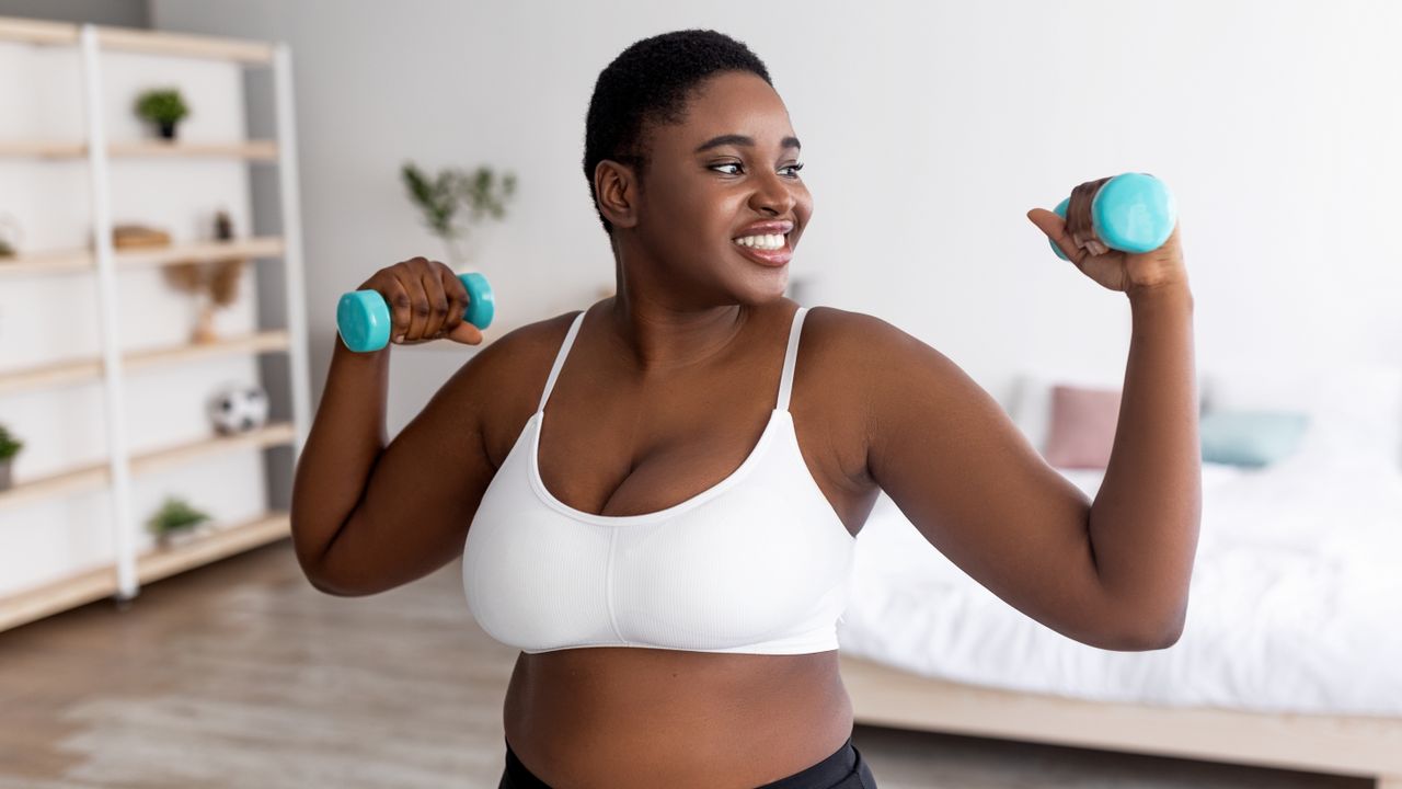 A woman stands in a bedroom holding light weights, with her elbows bent out to the sides and the weights held above shoulder height. She is smiling and we see a bed and decorative shelves behind her.