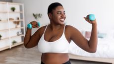 A woman stands in a bedroom holding light weights, with her elbows bent out to the sides and the weights held above shoulder height. She is smiling and we see a bed and decorative shelves behind her.