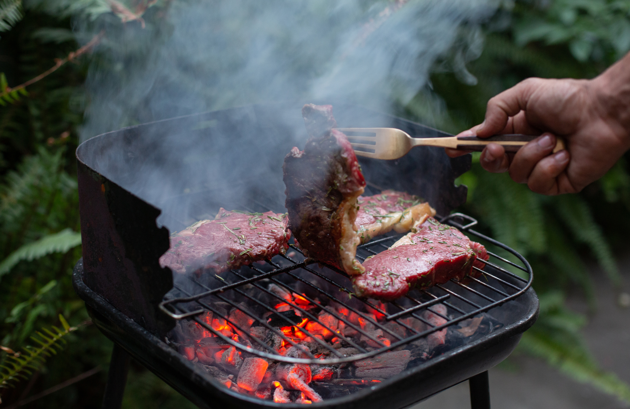 A person cooking steaks on a smoky BBQ in their garden.