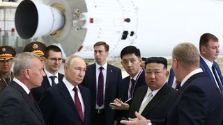 eight men in dark suits stand in front of a rocket inside a spaceport facility.