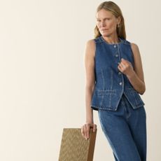 A woman leaning on the top of a wicker chair wearing a denim vest and jeans from Whistles