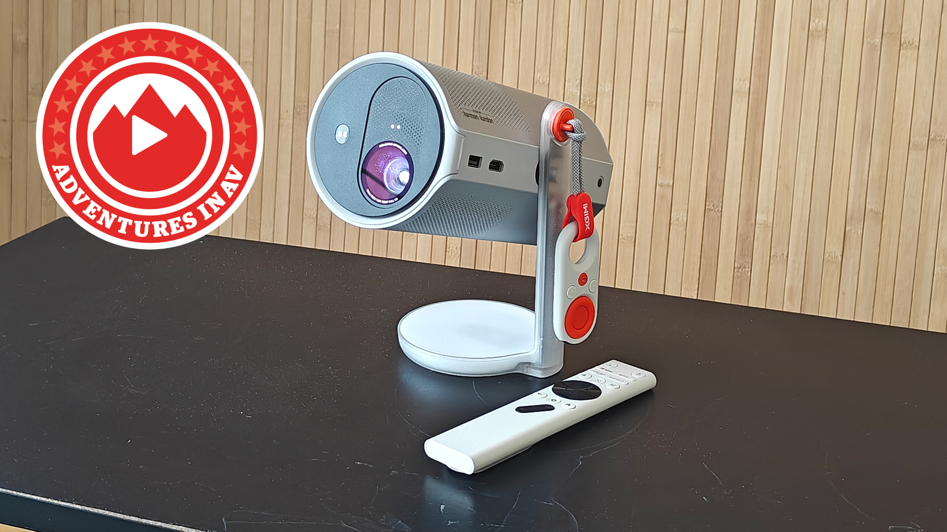 An Xgimi MoGo 4 projector on a black table with a wooden backdrop with an "Adventures in AV" logo to the left