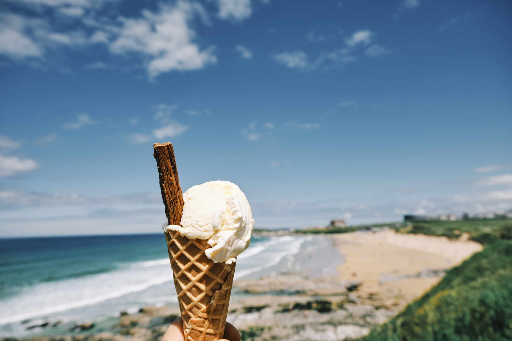 A person holding an ice cream with a flake, in front of a Cornwall beach view.