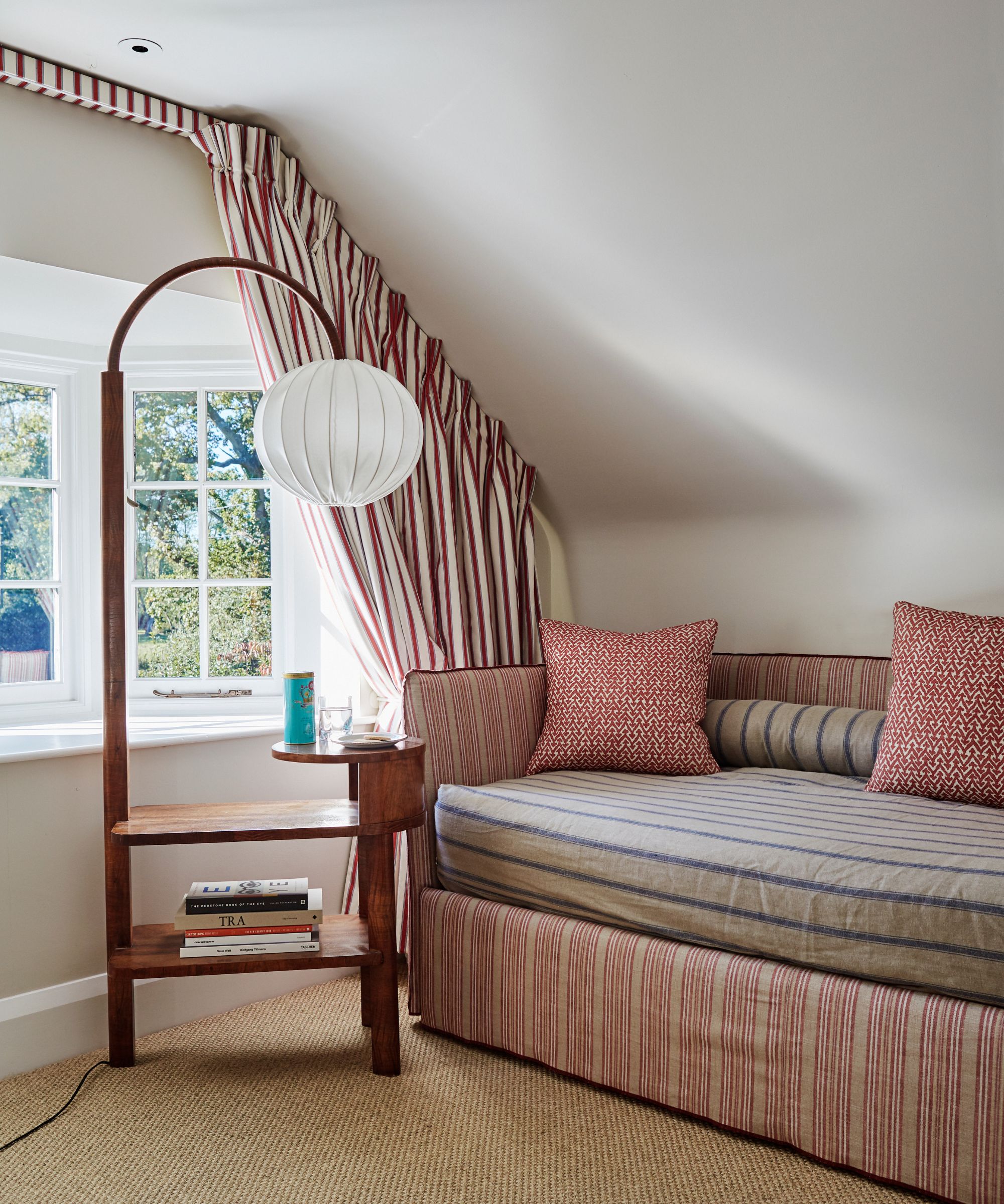 A white attic space with striped curtains, a striped sofa, and a brown floor lamp with a white lampshade in front of bay windows.
