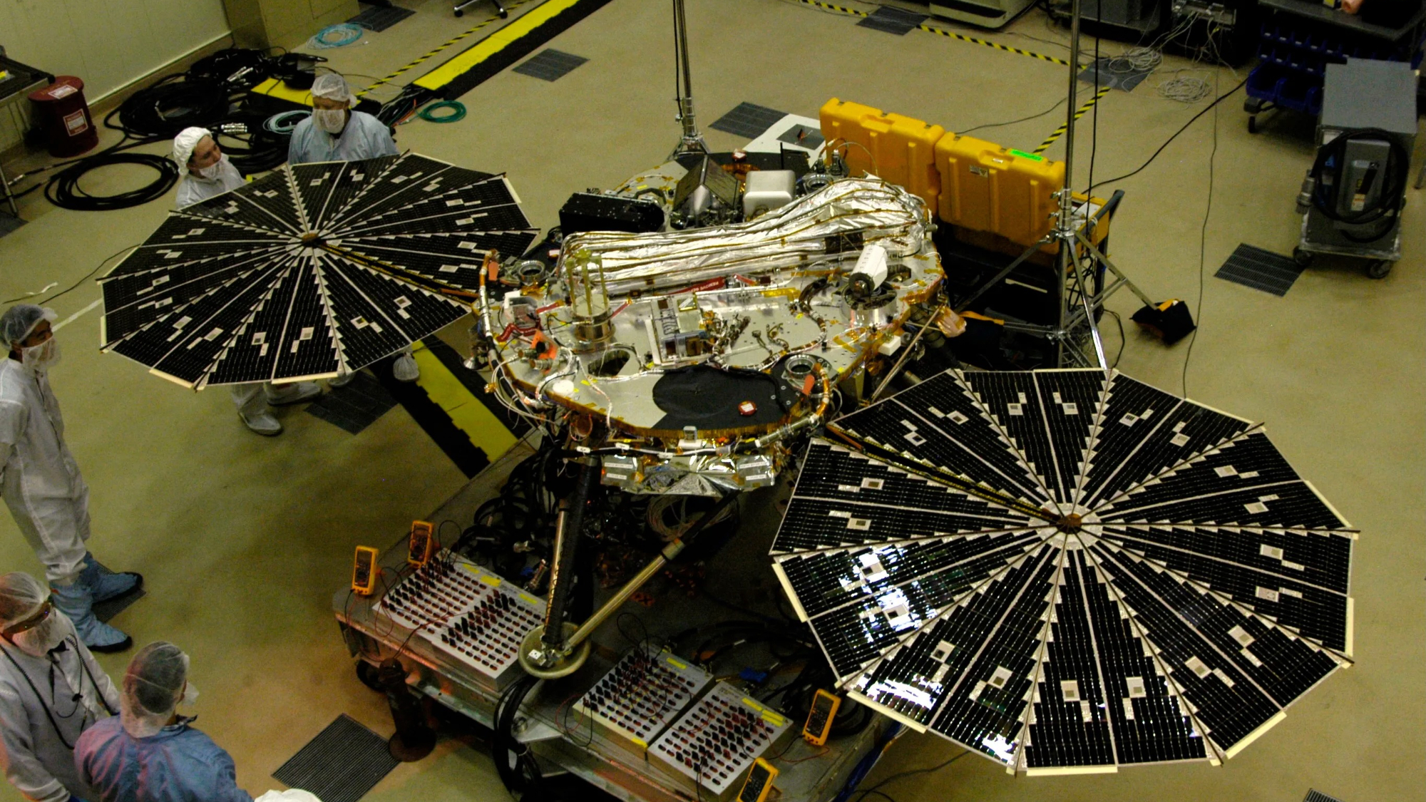 a circular silver spacecraft with twin circular solar arrays extended sits in a large clean room with five technicians nearby