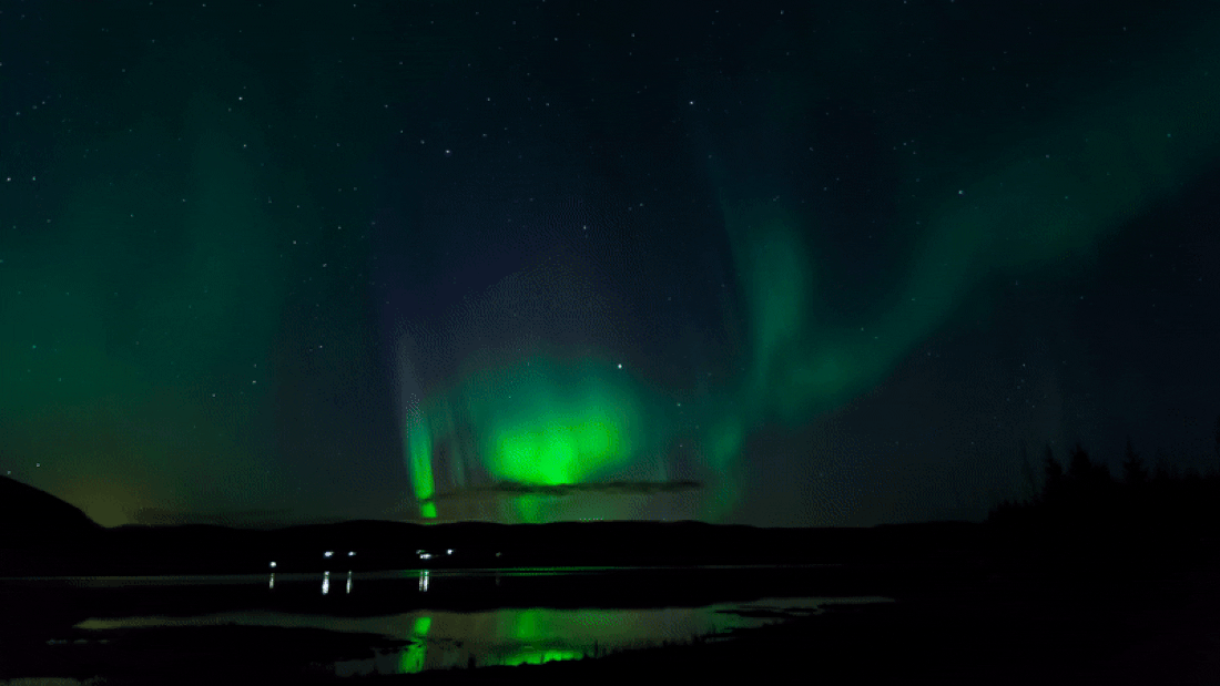 The Northern Lights glow greenly over a lake in Iceland, with the lights of a small town just visible on the other side.