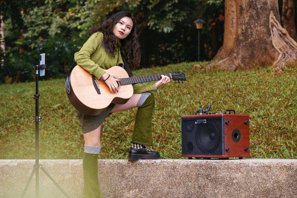 A woman playing guitar next to a Joyo BSK-80 with her phone, attached to a phonestand, in front of her