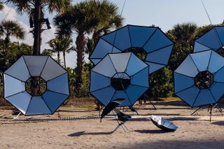 Daytime, Miami Beach, sand, cluster of silver solar reflectors on black frames, palm trees, blue cloudy sky, two small black umbrellas, one attached to a black beach chair