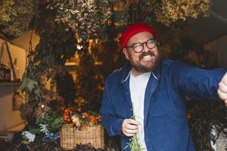 Banjo stands inside a small shed with flowers and herbs hanging from the walls and ceiling