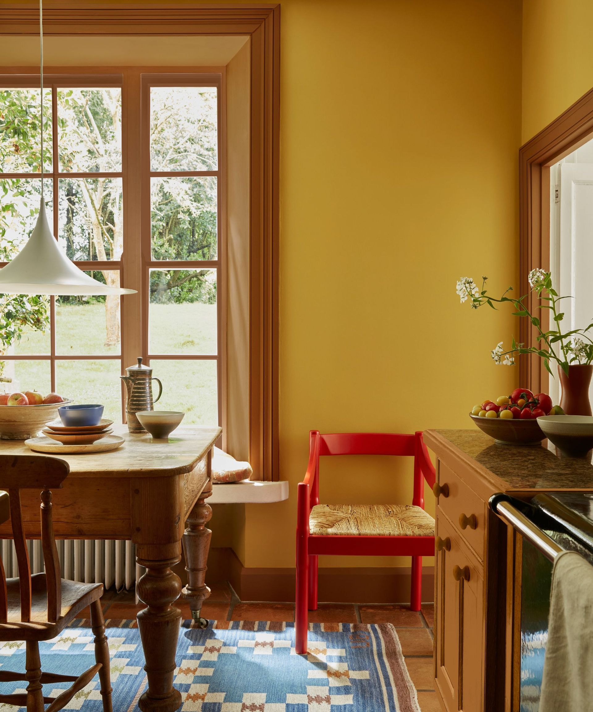 Yellow kitchen with blue woven rug, red painted chair, wood dining table and chairs and low pendant light