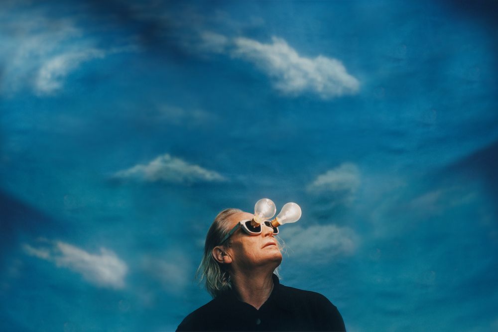 A man in a black shirt looking through glasses with lightbulbs attached to the lenses, against a blue sky with clouds