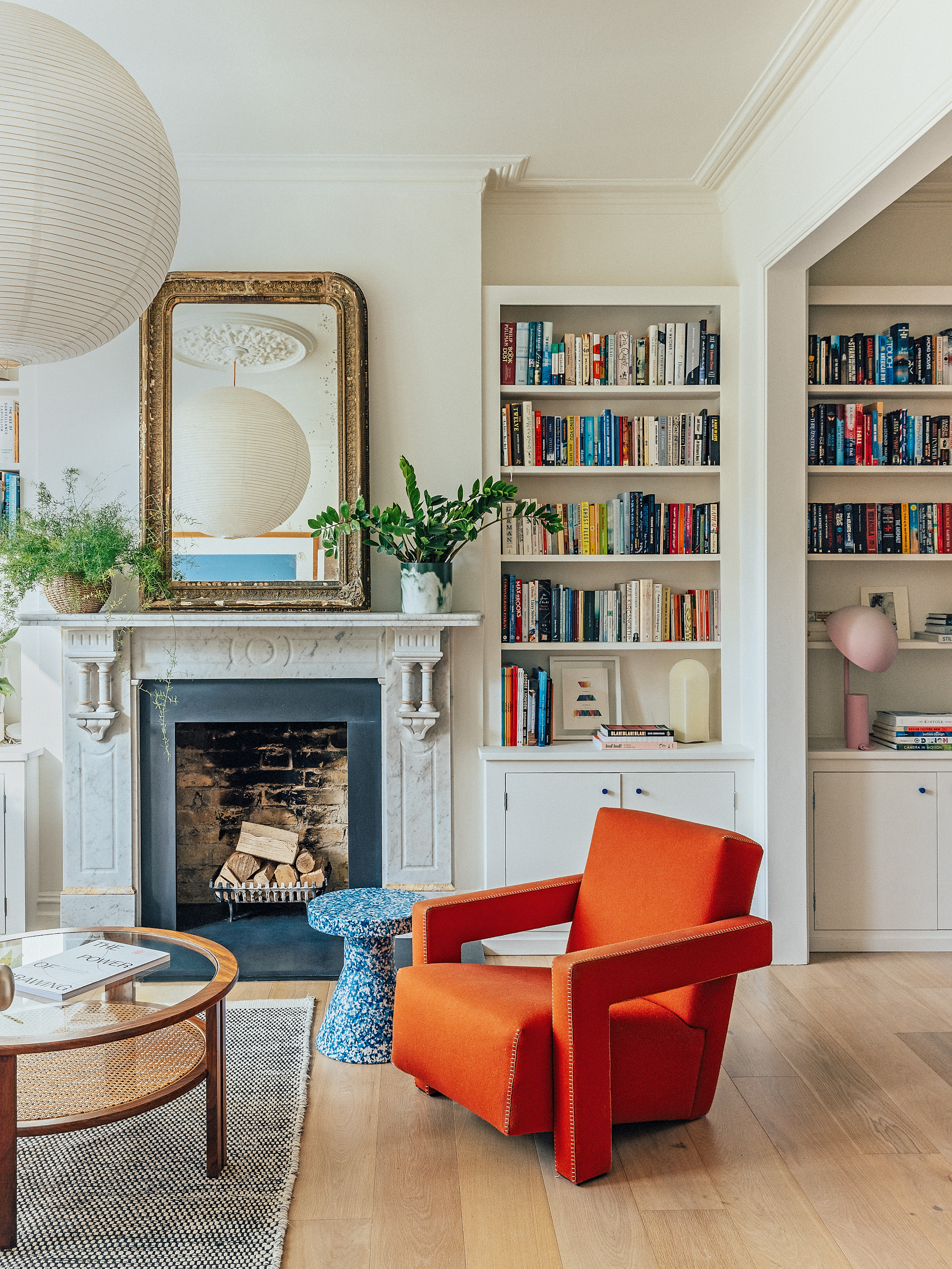 White living room with oak floor, white marble fireplace, built-in bookshelf, round wood, glass and cane table, orange armchair and blue speckled side table