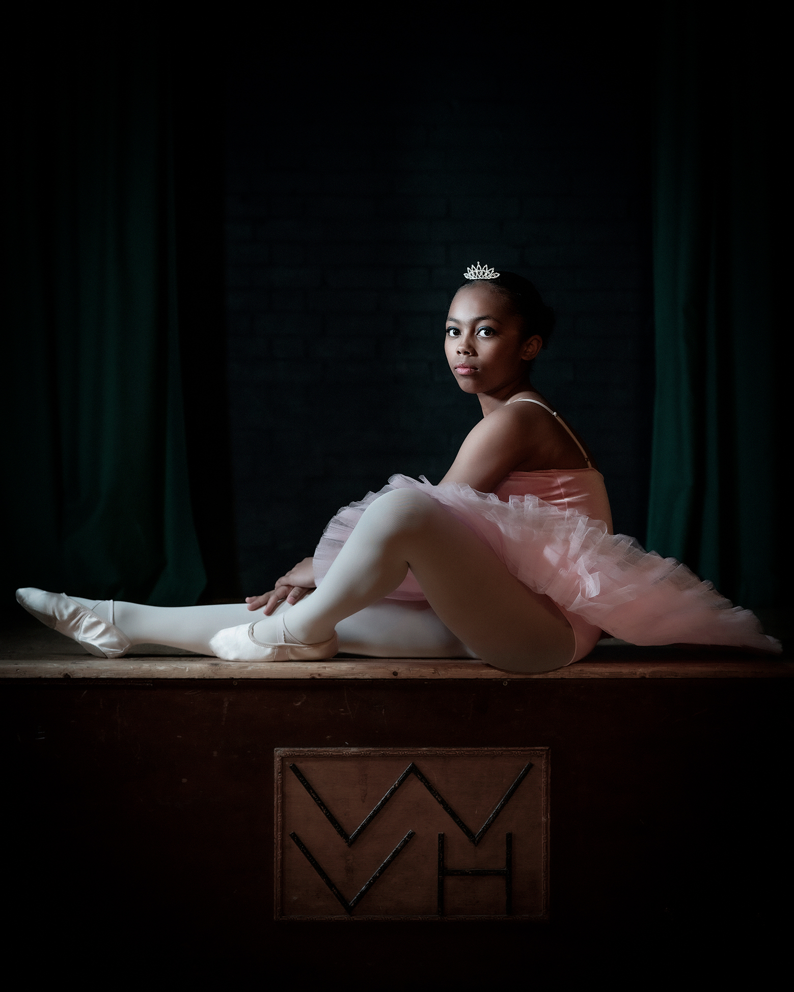 A ballet dancer poses gracefully in a pink tutu, sitting on a wooden platform, with dark curtains in the background