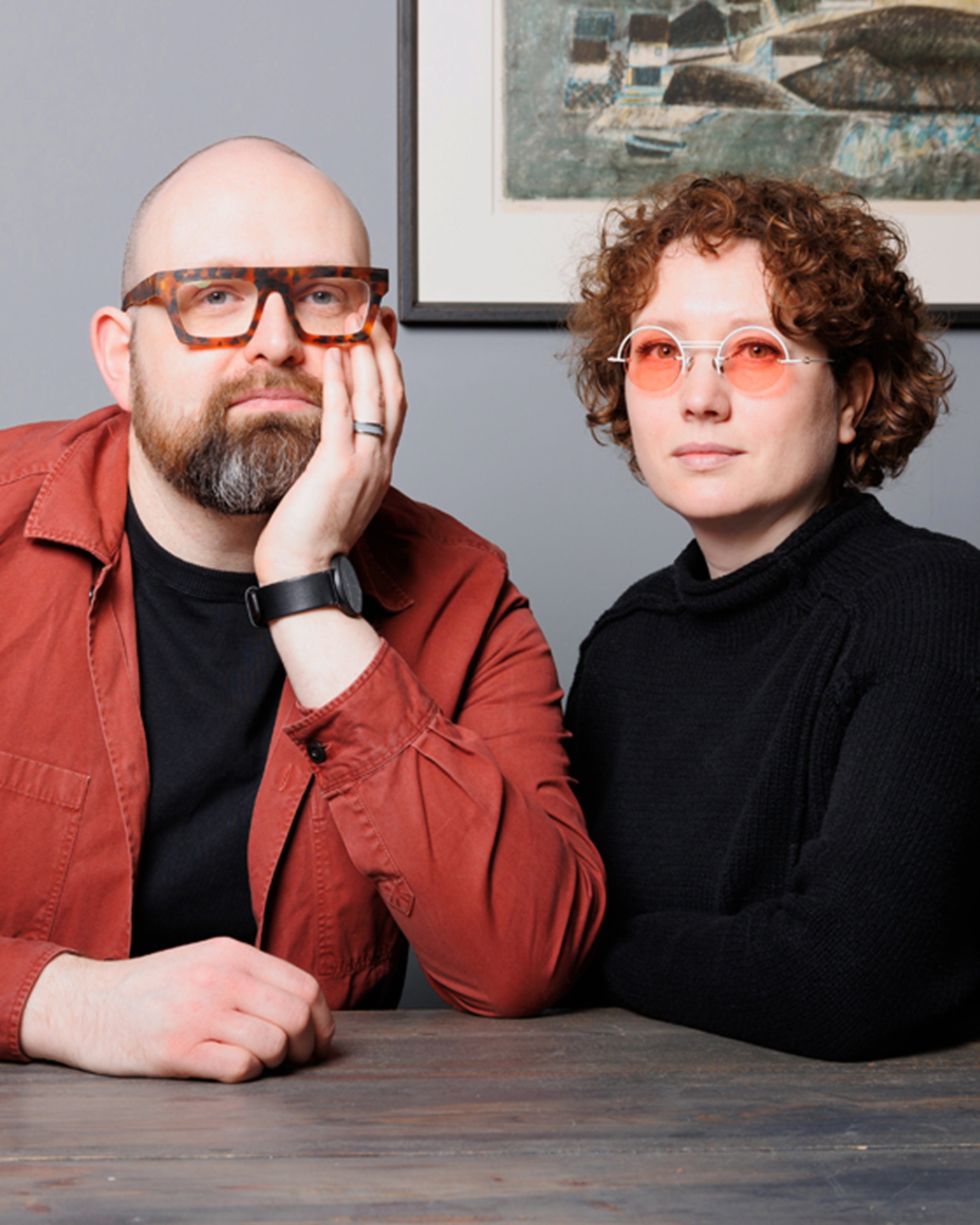Studio portrait of a man with a beard and bold tortoiseshell glasses leaning his head on his hand next to a woman with short red curly hair wearing pink-tinted glasses