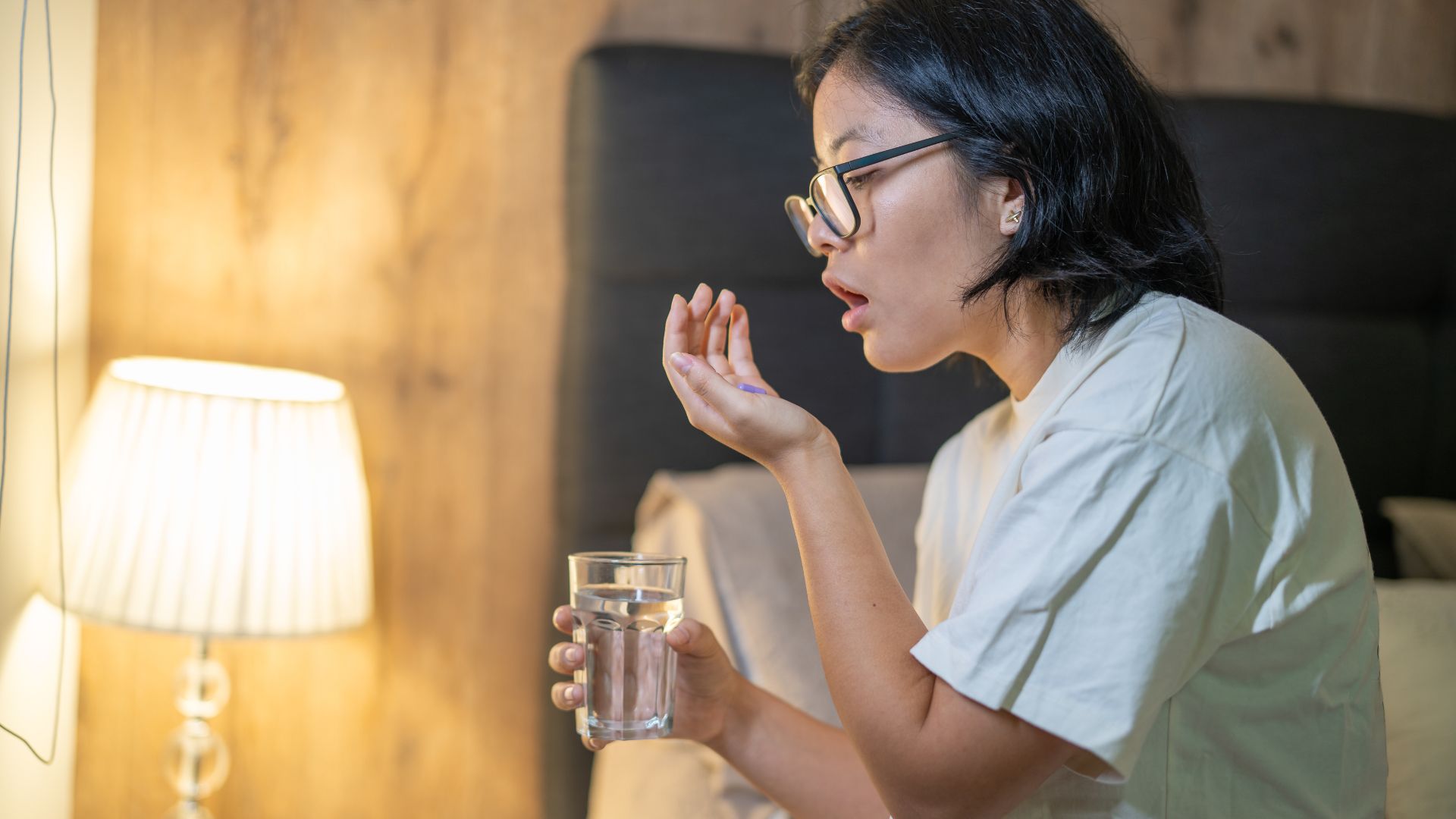 A woman with black hair wearing glasses and holding a glass of water sits on the edge of her bed and prepares to take a multivitamin supplement at night. 