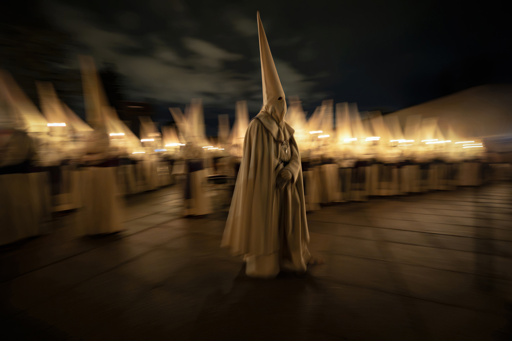 A hooded figure in white robes walks among a procession of similar figures, illuminated by flickering candles on a dark night